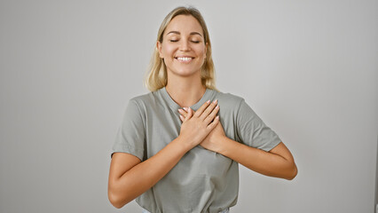 A contented young woman with closed eyes stands against a white background, hands over heart,...
