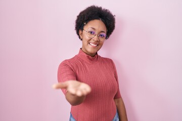 Beautiful african woman with curly hair standing over pink background smiling cheerful offering...