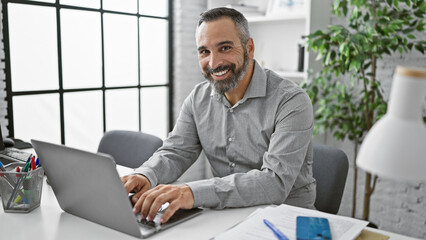 Smiling hispanic senior man with a grey beard working on a laptop in a modern office interior.