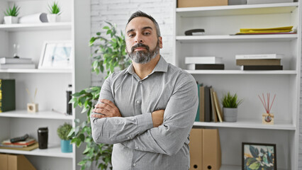 Confident mature hispanic man with a beard stands arms-crossed in a modern office setting.