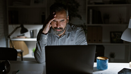 Mature hispanic man with grey beard stressing at night in dark office