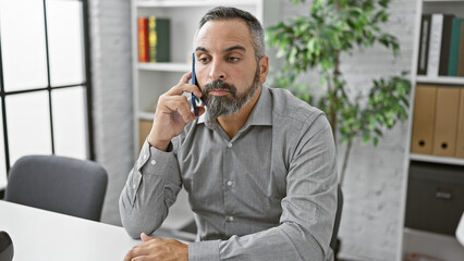 Handsome senior hispanic man with grey beard in an office setting talking on a smartphone