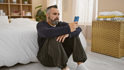 Mature bearded hispanic man sits in bedroom using smartphone, surrounded by neutral decor.