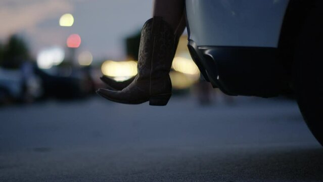 Woman wearing cowboy boots sits in the back of her car while tailgating at a country music concert