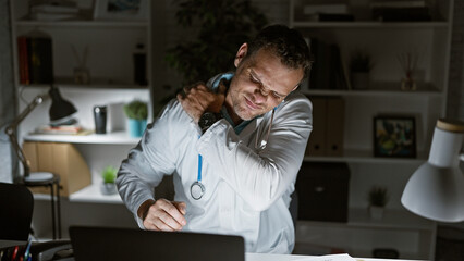 Hispanic male doctor feeling neck pain during a late night at the office, portraying workplace...