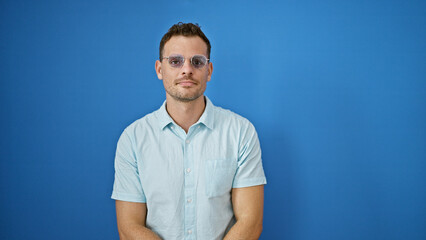 A young adult hispanic man in a casual shirt and sunglasses stands before a plain blue wall...