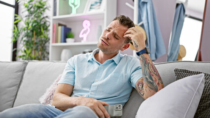 Concerned tattooed man sitting on couch at home with phone in hand, looking stressed.
