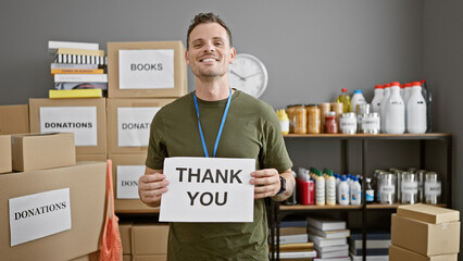 A smiling man holding a 'thank you' sign surrounded by donation boxes in an indoor charity center.