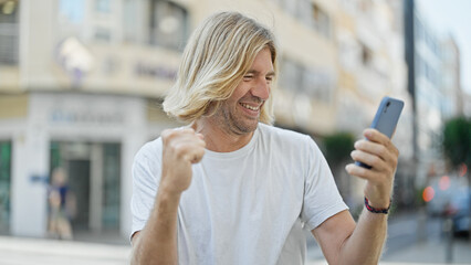 Handsome young man celebrating with a fist pump while looking at his smartphone on a city street.