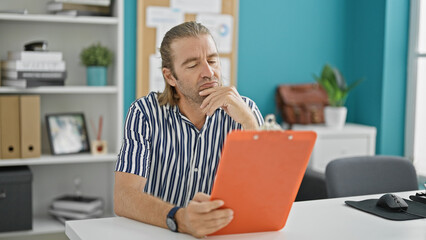 Pensive man with long blond hair contemplating a clipboard in a modern office setting