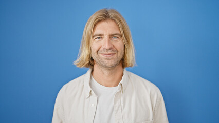 Handsome adult man with blond long hair smiling against a plain blue background.