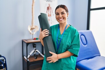 Young beautiful hispanic woman physiotherapist smiling confident holding yoga mat at rehab clinic