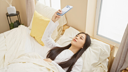 Obraz premium A young asian woman relaxes in a bedroom, taking a selfie with her smartphone, exemplifying modern indoor lifestyle.