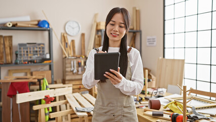 Asian woman using tablet in a woodworking workshop full of tools and timber.