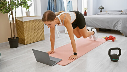 A focused young woman exercises in her bedroom with a laptop and fitness equipment on the wooden...
