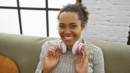 A cheerful woman with curly hair enjoying music with pink headphones in a cozy living room.