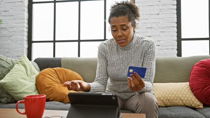 A perplexed woman holding a card sits on a sofa, using a tablet in a cozy living room.