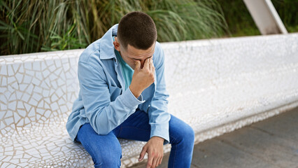A young hispanic man in casual attire appears stressed while sitting on a park bench outdoors.