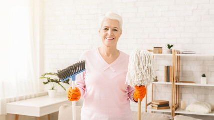 Senior woman standing while holding a mop and a brush in her hands. She appears to be ready to...