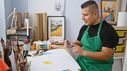 Young, confident latin artist guy making smartphone photos of his art indoors, ready to draw a...