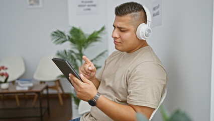 Relaxed yet serious young latin man engrossed in his internet lifestyle, concentrating on touchpad screen and listening through headphones while sitting casual in waiting room chair