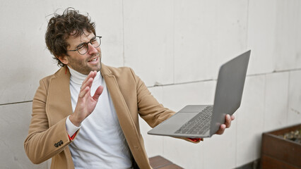 Young hispanic man engaging in a video call on a laptop while standing on an urban street.
