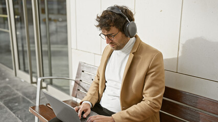 Handsome young hispanic man with beard and headphones using laptop on city street bench.