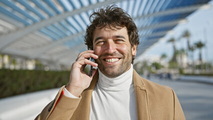 Handsome young man with beard smiling during a phone call in a green city park outdoors.