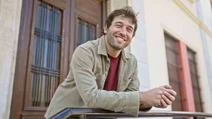 Smiling, bearded hispanic man leaning on a railing with a blurred building facade in an urban...