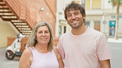 Confident mother and son standing together on a sunny city street, their smiles radiating with...