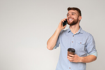 Young bearded man holding cup of coffee and talking on phone on beige background, panorama, copy...