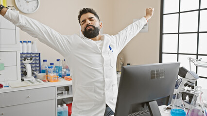 A young hispanic man stretches in a modern laboratory setting, showing relief or a break during his...