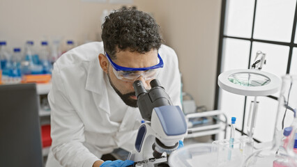 Hispanic scientist man uses microscope in laboratory research setting, wearing lab coat and protective goggles.