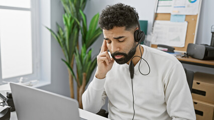 A focused hispanic man working indoors at an office with a laptop, wearing headphones and a headset.