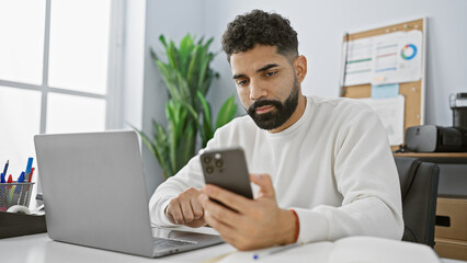 Hispanic man with beard using smartphone at office desk with laptop and indoor plant.