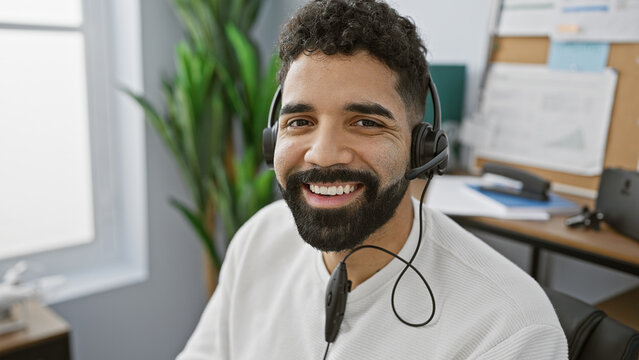 A smiling hispanic man wearing a headset in an office setting with a computer and plants in the background.