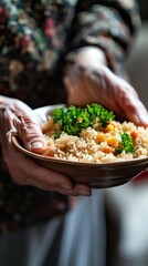 A close up of an elderly woman's hands holding a bowl of rice. The rice is topped with parsley. The woman is wearing a floral apron.