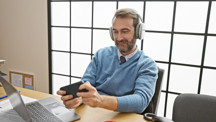 Handsome middle-aged hispanic man in a blue sweater using smartphone with headphones in office.