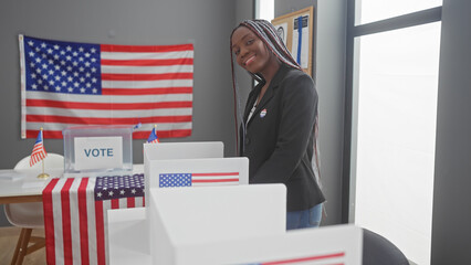 African american woman with braids smiles inside a voting center adorned with united states flags.