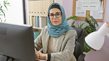 Professional middle-aged woman wearing a hijab and glasses sits at her office workstation in front...