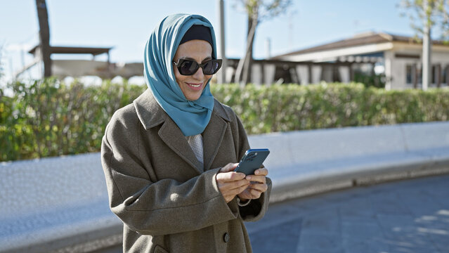 A smiling mature woman in a hijab using a smartphone in a sunny park - Powered by Adobe