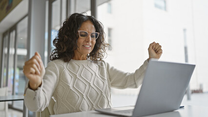 An exuberant hispanic middle-aged woman celebrates a success at her office workplace in front of a laptop.