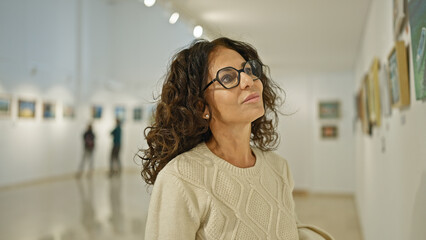 Mature hispanic woman reflects thoughtfully in a bright art gallery setting, embodying culture,...