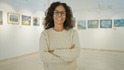 A smiling, mature hispanic woman with crossed arms stands confidently in a bright art gallery.