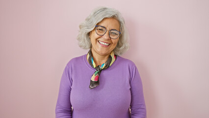 A cheerful mature woman with grey hair, glasses, and wearing a purple sweater, poses against a pink...