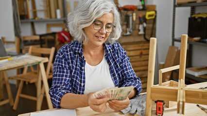 A grey-haired woman counts polish zloty bills in a carpentry workshop, surrounded by woodworking...