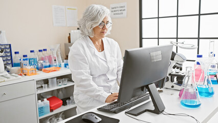 Mature woman scientist working on computer in laboratory setting with chemicals and microscope