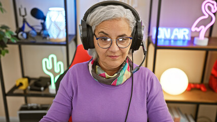 Mature woman with headphones in a colorful gaming room looks at camera