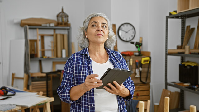 Thoughtful mature woman using tablet in a well-organized carpentry workshop, reflecting creativity and craftsmanship.