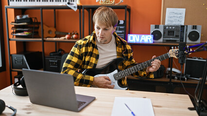 A focused blond man rehearses on an electric guitar in a music studio with recording equipment.
